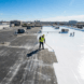 Professional roofer applying bright white silicone roof coating to a weathered EPDM rubber membrane.