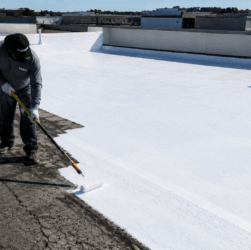 Professional roofer applying bright white silicone roof coating to a weathered EPDM rubber membrane.