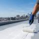 A professional roofer applies a white elastomeric coating to seal a seam on a flat TPO roof to prevent leaks.